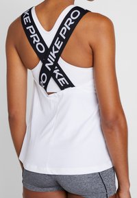 Top deportivo blanco sin mangas con tirantes con el logo negro "NIKE PRO", que presenta un corte relajado y un diseño con escote en la parte posterior. Pantalones cortos grises visibles por debajo.