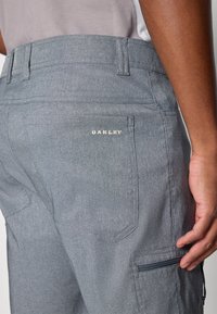 Calças cinzentas Oakley com um tecido texturizado, apresentando um bolso lateral com fecho e um logótipo bordado na parte de trás. Ajuste bem talhado e design estruturado.