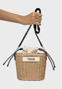 Borsa a secchiello in paglia intrecciata con chiusura a cordoncino beige, manici neri e etichetta crema con la scritta "TOUS." Superficie texturizzata con fibre naturali.
