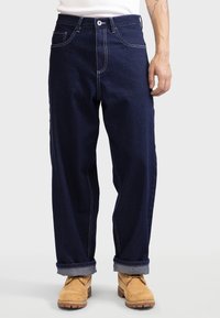 Mann in dunkelblauen, umgeschlagenen Jeans und braunen Leder-Arbeitsstiefeln, der vor einem einfachen hellen Hintergrund steht.