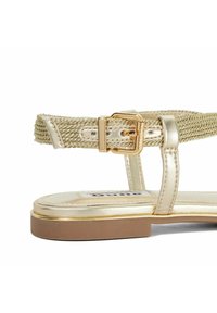 Gouden metallic platte sandaal met een geweven band en een gespsluiting, een glad leren bovenwerk en een contrasterende bruine zool.