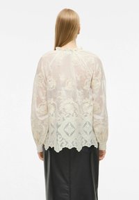 Lys beige broderet bluse med lange ærmer, præget af blomsterdesigns og en scallop-kant. Gennemsigtigt materiale med flæser i kraven.
