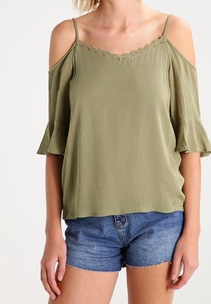 Femme portant un chemisier vert olive à épaules dénudées avec bordure en dentelle et un short en jean bleu sur un fond uni.