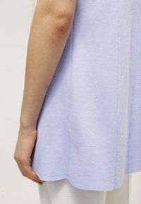 Ljuslila topp med korta ärmar, tillverkad av mjukt tyg. De nedre kanterna har en lätt kurva och synliga sömdetaljer.