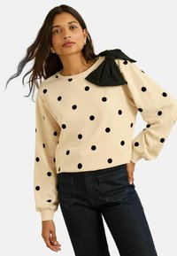 Béžový sveter s čiernymi bodkami, na ktorom sa nachádza veľká čierna mašľa na ramene. Dlhé rukávy, okrúhly výstrih a voľný strih.
