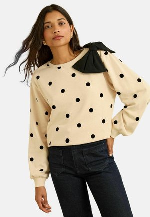 Béžový sveter s čiernymi bodkami, na ktorom sa nachádza veľká čierna mašľa na ramene. Dlhé rukávy, okrúhly výstrih a voľný strih.