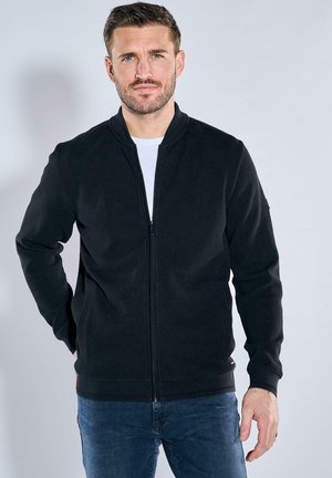 Black zip-up jacket with a textured surface, stand collar, and two side pockets, paired with a white t-shirt and blue jeans.