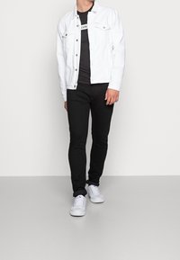Muž v bílé džínsové bundě, černém tričku s potiskem, černých slim-fit kalhotách a bílých teniskách stojí na jednobarevném pozadí.