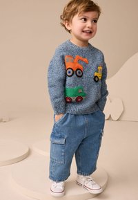 Blauer gestrickter Pullover mit orangefarbenen und grünen Lkw-Applikationen, kombiniert mit hellen Cargo-Jeans und weißen Turnschuhen.