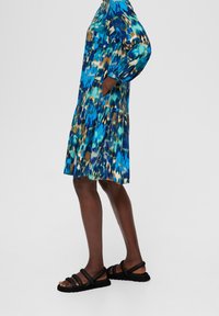 Blau und grün gemustertes Kleid mit langen Ärmeln, schichtweissem Saum und Seitentaschen, kombiniert mit schwarzen Riemchensandalen.