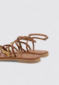 Sandalias de cuero marrón con múltiples correas y una hebilla dorada. Tiene una correa trenzada en colores vibrantes. Suelas planas con una textura suave.