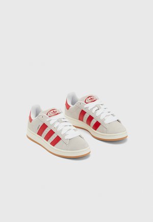 Paar beige Sneaker mit roten Streifen und Fersenakzenten, weißen Schnürsenkeln und gummifarbener Sohle auf einfachem hellem Hintergrund.