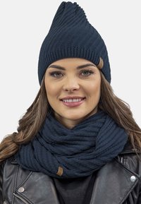 Caciula tricotată în nuanță de bleumarin, cu un design texturat în zigzag, având un vârf ascuțit. Asortată cu o eșarfă cu nervuri, tot în nuanță de bleumarin.