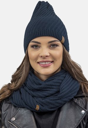 Caciula tricotată în nuanță de bleumarin, cu un design texturat în zigzag, având un vârf ascuțit. Asortată cu o eșarfă cu nervuri, tot în nuanță de bleumarin.