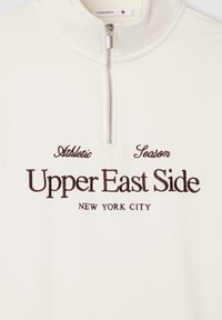 Biała polarowa bluza z krótkim zamkiem na szyi z haftowanym tekstem w ciemnym brązie: "Athletic Season Upper East Side Nowy Jork." Gładka faktura.