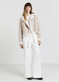 Giacca in finta suede beige con rifiniture in shearling bianco, chiusura con zip e design leggermente cropped. Abbinata a pantaloni bianchi a gamba larga.