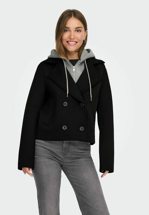 Smilende kvinde iført en sort dobbeltradet frakke over en grå hættetrøje og grå jeans, stående foran en ensfarvet hvid baggrund.