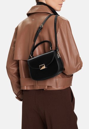 Frau trägt braune Lederjacke und dunkle Hose, mit einer schwarzen Ledertasche mit goldener Schnalle über der Schulter.