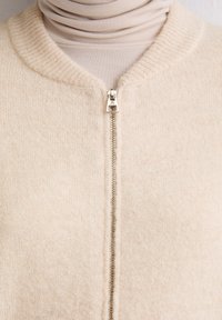 Cardigan a zip con zip color crema realizzato in morbido materiale, con un colletto a costine e una cerniera argentata. La texture appare accogliente e liscia.