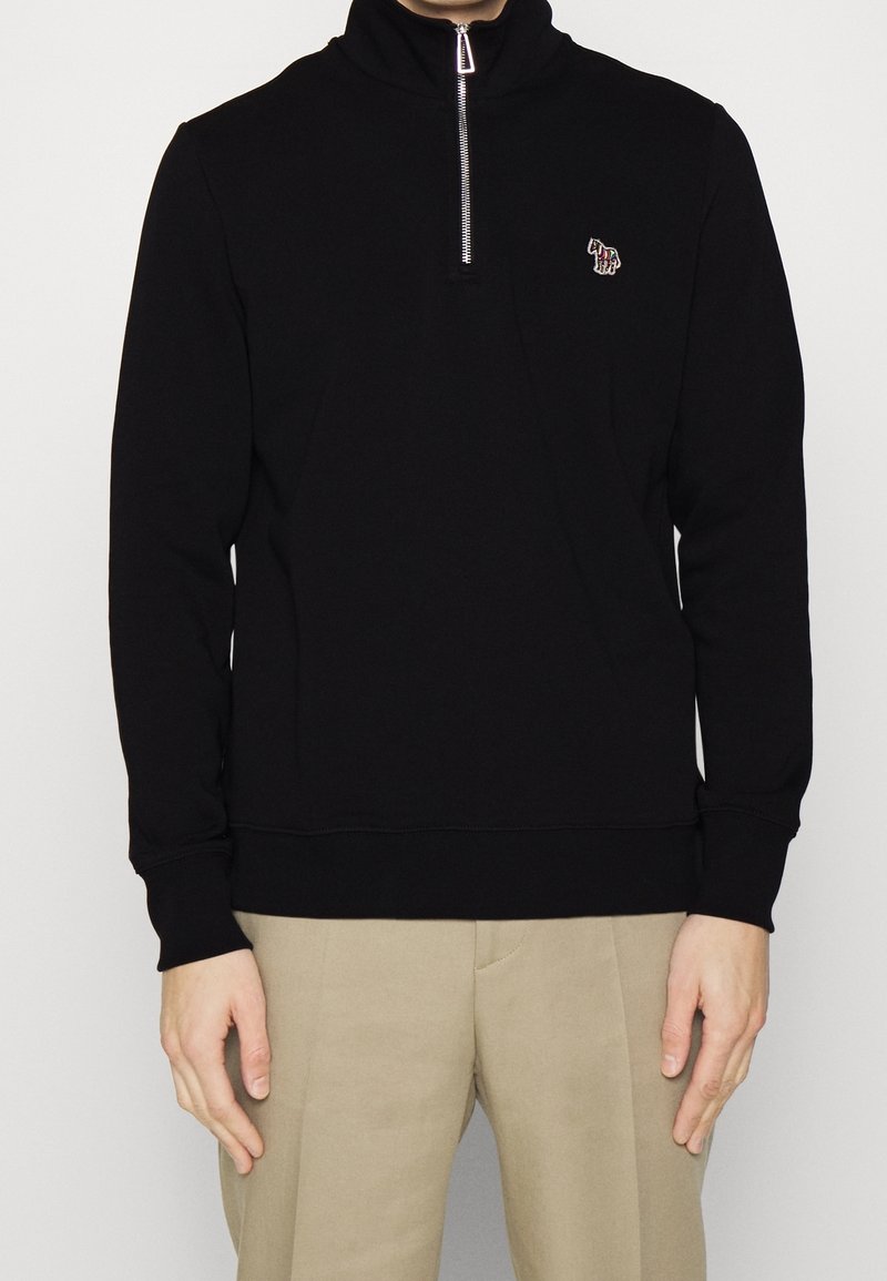 Personne portant un sweat-shirt noir à zip quart avec un petit logo zèbre coloré sur la poitrine et un pantalon beige sur un fond uni.