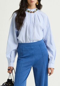 Blusa a maniche lunghe a righe blu e bianche con maniche a sbuffo, caratterizzata da un colletto alto e una texture liscia. Indossata con pantaloni blu texturizzati.