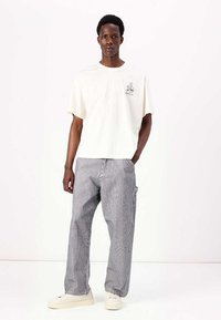 Camiseta blanca de manga corta con un pequeño gráfico, combinada con pantalones anchos a rayas en tonos de azul y gris, y zapatos de colores claros.