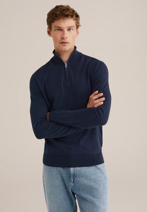 Un jeune homme aux cheveux bouclés portant un pull zippé bleu marine et un jean bleu clair, se tenant les bras croisés devant un fond neutre.