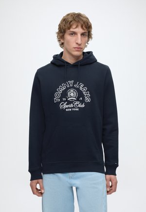 Młody mężczyzna ubrany w granatową bluzę z kapturem Tommy Jeans Sports Club i jasnoniebieskie dżinsy, stojący na jednolitym białym tle.