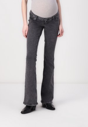 Kobieta w ciąży ubrana w szare elastyczne jeansy ciążowe i czarne botki na obcasie, stojąca na białym tle.