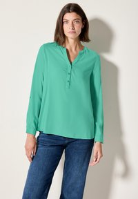 Blouse turquoise à manches longues avec patte de boutonnage, coupe décontractée, ourlet arrondi et texture de tissu douce, associée à un jean bleu pour le contraste.