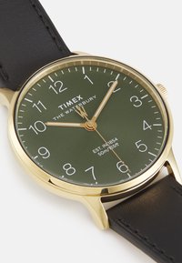 Wodoszczelny zegarek Timex z zieloną tarczą, złotą kopertą i czarnym skórzanym paskiem. Białe wskaźniki godzinowe i wskazówki z złotymi akcentami.