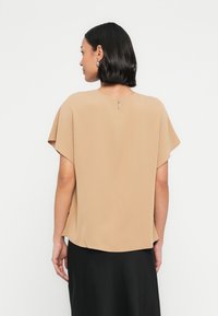 Μπεζ μπ�λούζα με κοντά μανίκια από ελαφρύ ύφασμα, με στρογγυλεμένο λαιμόκοψη στο πίσω μέρος και λεπτομέρεια φερμουάρ, με χαλαρή σιλουέτα.