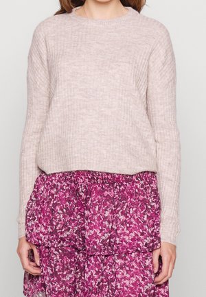 Femme portant un pull côtelé beige clair et une jupe fleurie rose superposée, debout devant un fond clair uni.