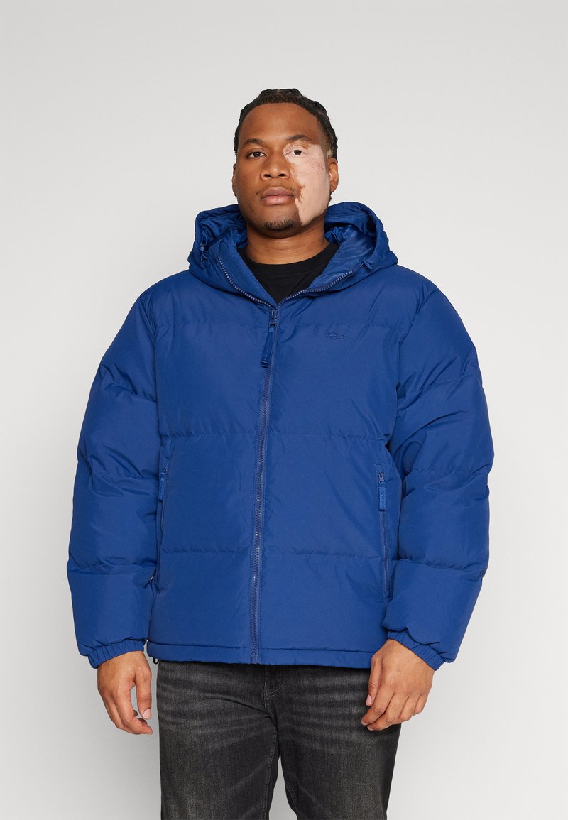 Giacca a vento blu con zip frontale, cappuccio regolabile e polsini elastici. Materiale liscio con una finitura leggermente lucida.