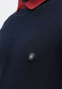 Personne portant un pull en tricot bleu marine sur une chemise rouge à col, avec un petit emblème rond noir présentant des lettres argentées entrelacées sur la poitrine.