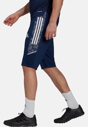 Mężczyzna idący w granatowych szortach sportowych Adidas z białymi paskami i wzorami, w białych skarpetkach i czarnych butach sportowych.
