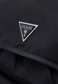 Črn najlon nahrbtnik z usnjeno srebrno trikotno oznako z besedilom "GUESS USA" in vprašajem, ki je podrobno obdelan z teksturiranim zaključkom.