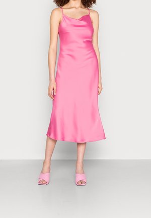Kobieta w różowej satynowej sukience midi na ramiączkach z cienkimi paskami oraz pasujących różowych sandałach na słupku z odkrytymi palcami.