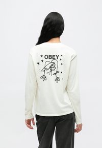 Crèmekleurig T-shirt met lange mouwen, voorzien van een zwart gedrukte afbeelding, het woord "OBEY" en een lijntekening met sterren op de achterkant.