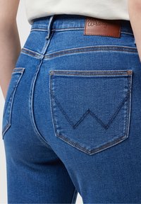 Los jeans de mezclilla azul de tiro alto presentan una etiqueta de marca de cuero marrón y un diseño distintivo en el bolsillo trasero con un patrón de costura en forma de chevrón.