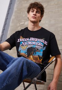 Black cotton t-shirt featuring a colorful graphic print of butterflies and a figure in a field, with "DAYS BEFORE SUMMER" in pink text.