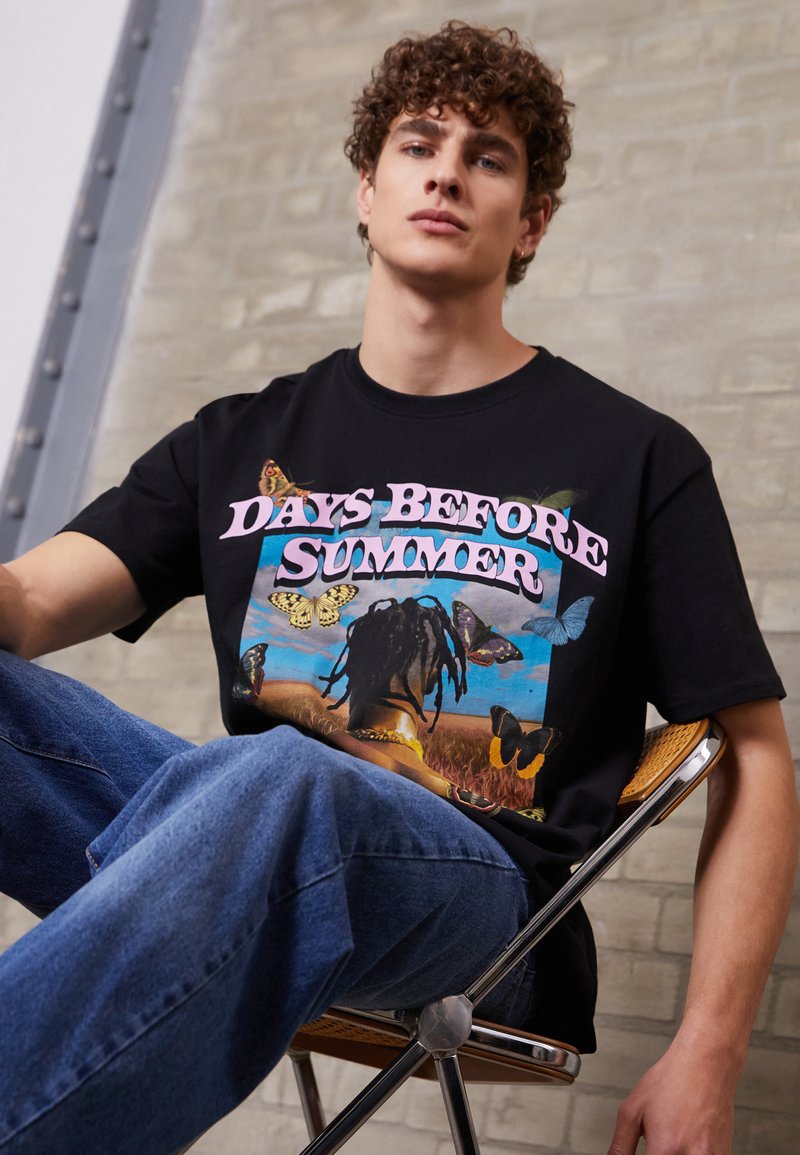 Black cotton t-shirt featuring a colorful graphic print of butterflies and a figure in a field, with "DAYS BEFORE SUMMER" in pink text.
