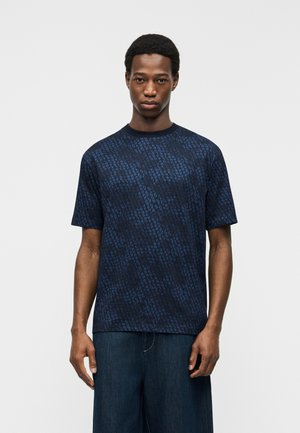 Jeune homme portant une chemise à manches courtes bleu foncé à motifs et un pantalon ample bleu foncé, debout devant un fond clair uni.