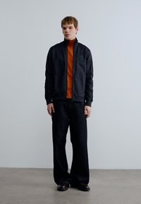 Μαύρο μπουφάν με φερμουάρ πάνω από πορτοκαλί ζιβάγκο, συνδυασμένο με ευρύχωρο σκούρο παντελόνι και μαύρα παπούτσια. Λιτή υφή, μοντέρνο σχέδιο.