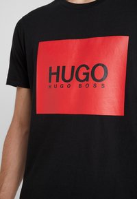 Černé tričko s červeným obdélníkovým grafickým prvkem, na kterém je výrazným černým písmem nápis "HUGO" a pod ním menším písmem "HUGO BOSS".