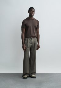 Mann steht vor einer schlichten Wand und trägt ein eng anliegendes braunes Kurzarmshirt, weit geschnittene graue Hose mit Gürtel und beige Schuhe.