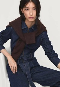 Combinaison en denim foncé à manches longues, fermeture éclair, et taille ceinturée, associée à un pull en tricot brun drapé sur les épaules.