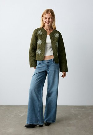 Donna sorridente che indossa una giacca trapuntata verde oliva con toppe floreali, top corto bianco, jeans a gambe larghe e scarpe nere, in piedi all'interno.