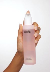 Rosa glasflaska med ett vitt pump-lock, märkt "BABOR SOOTHING ROSE TONER" i svart text, hålls av en hand mot en vit bakgrund.