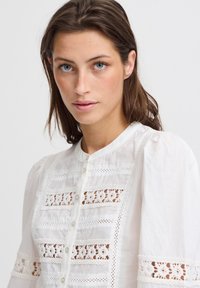 Blusa bianca con maniche a sbuffo, caratterizzata da pannelli in pizzo ricamato, chiusura con bottoni e delicate decorazioni in pizzo lungo i polsini.